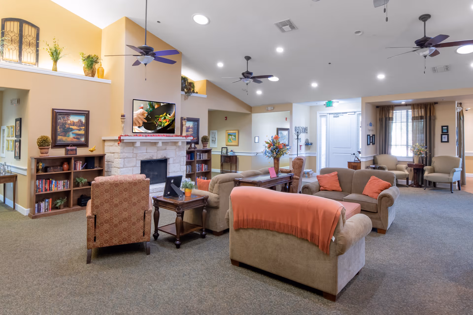 A spacious and well-lit living room area in an assisted living facility featuring comfortable sofas and armchairs arranged around a stone fireplace with a TV mounted above it. The room has ceiling fans, framed artwork on the walls, bookshelves filled with books, and decorative plants. Large windows with curtains allow natural light to enter the space.