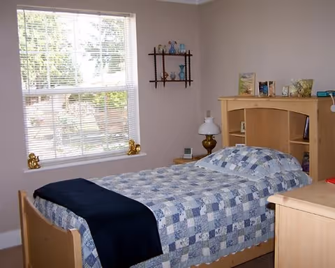 A small bedroom with a single bed covered in a blue and white patchwork quilt and a dark blue blanket at the foot. The bed has a light wood headboard with shelves holding books and decorative items. There is a window with white blinds letting in natural light, and small decorative figurines on the windowsill. A small wall shelf with glass vases is mounted on the wall beside the bed. The walls are painted a light beige color.