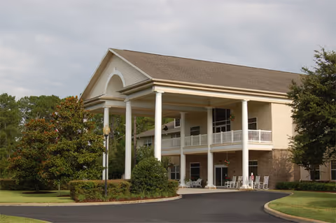 Exterior view of a senior living facility building with a large covered entrance supported by white columns, surrounded by trees and landscaping under a cloudy sky.