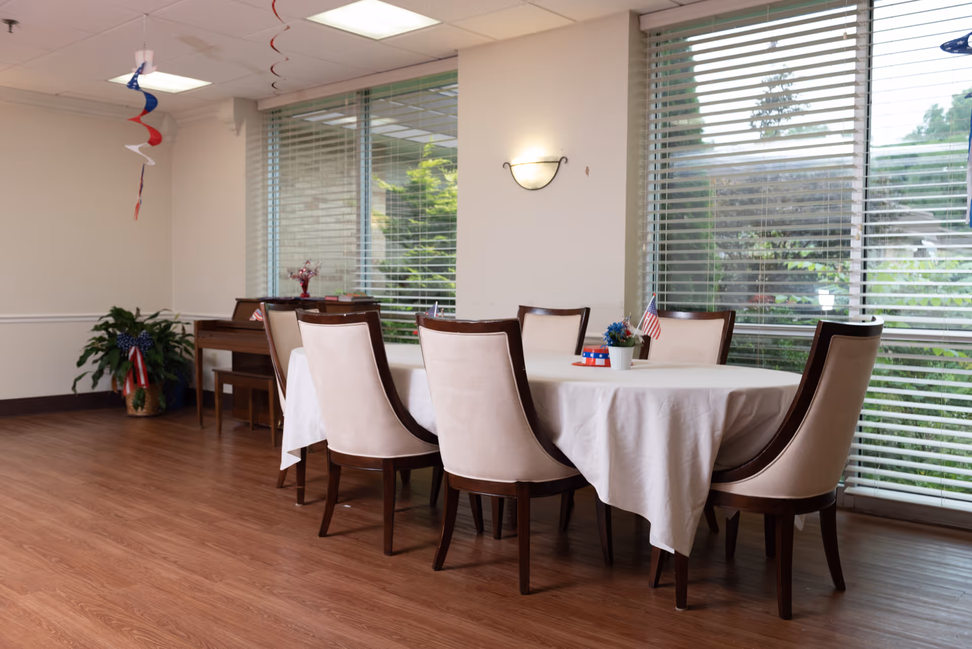 A dining room with a round table covered with a white tablecloth surrounded by eight beige upholstered chairs with dark wooden legs. The table has small patriotic decorations including a small American flag and a flower pot. Large windows with blinds allow natural light to enter the room. A piano and a plant with a small American flag are visible in the background. Red, white, and blue hanging decorations are suspended from the ceiling.
