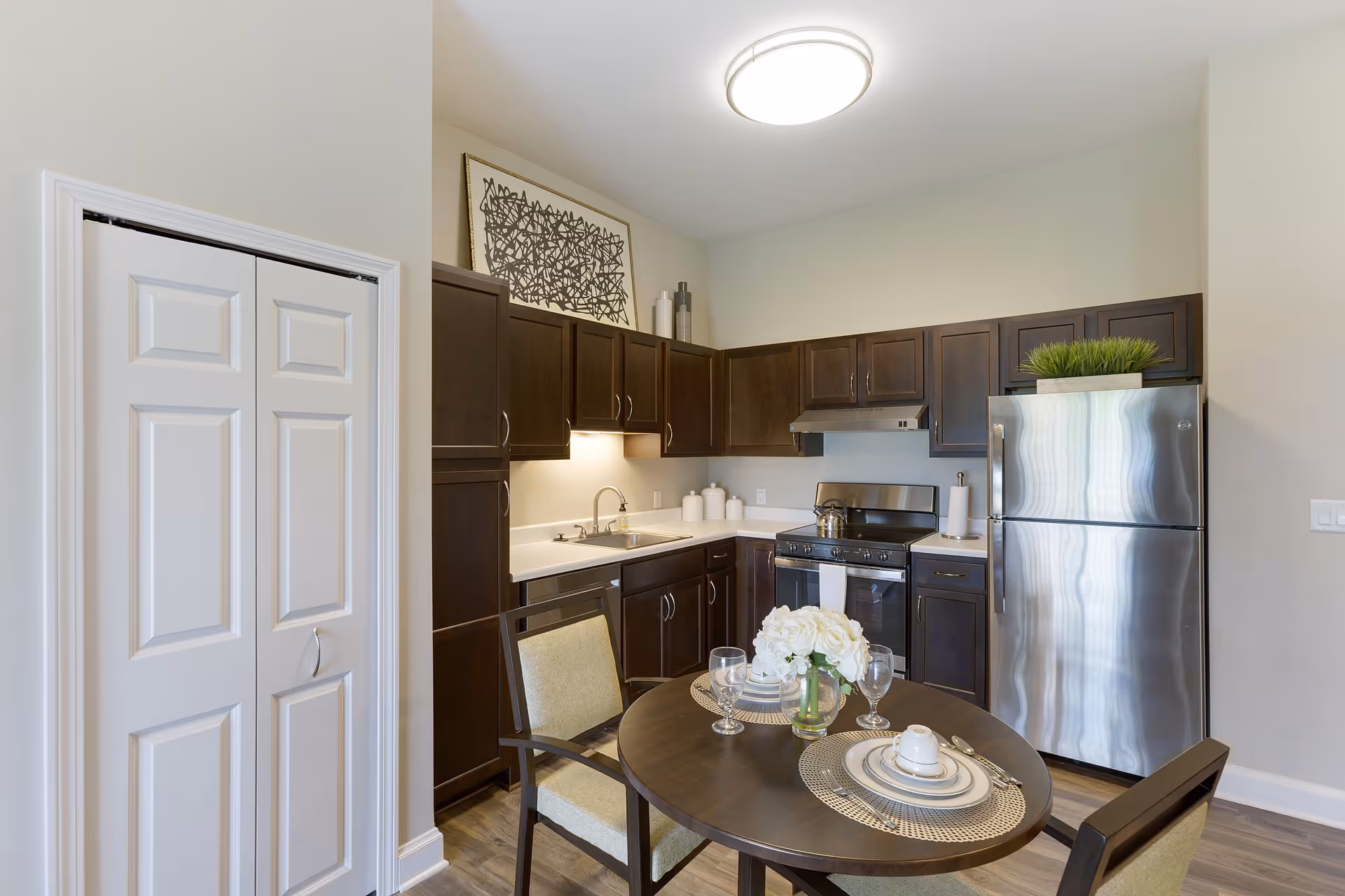 A modern kitchen with dark wood cabinets, a stainless steel refrigerator, stove, and sink. A round dining table with two chairs is set with plates, glasses, and a vase of white flowers. There is a piece of abstract art above the cabinets and a bright ceiling light.