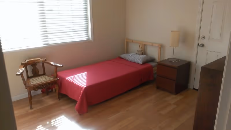 Small bedroom with a single bed covered by a red blanket, a wooden chair, nightstand with lamp, window blinds, and hardwood floor.