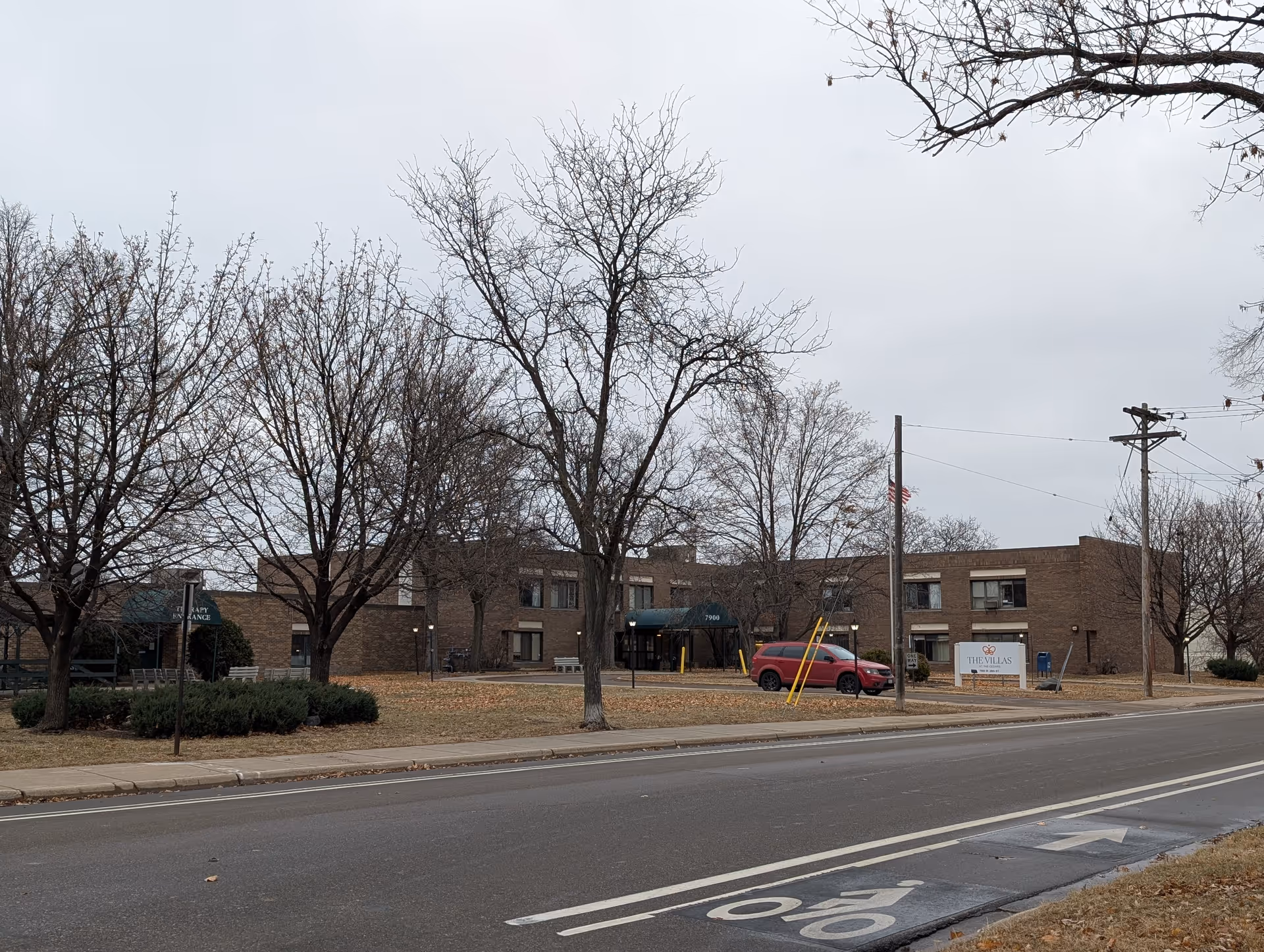 Exterior view of The Villas at The Cedars, a two-story brick building with leafless trees and a red SUV parked near the entrance. There is a sign in front of the building displaying the facility's name.