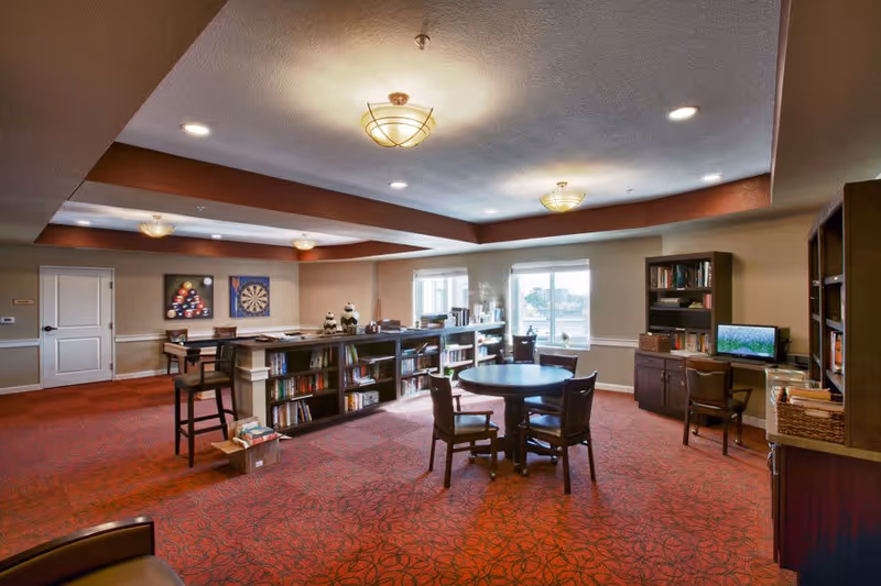 A spacious senior living common area with red patterned carpet, round table with four chairs, bookshelves filled with books, a computer desk with a monitor, and wall decorations including a dartboard and a billiards painting. The room is well-lit with ceiling lights and has two windows letting in natural light.