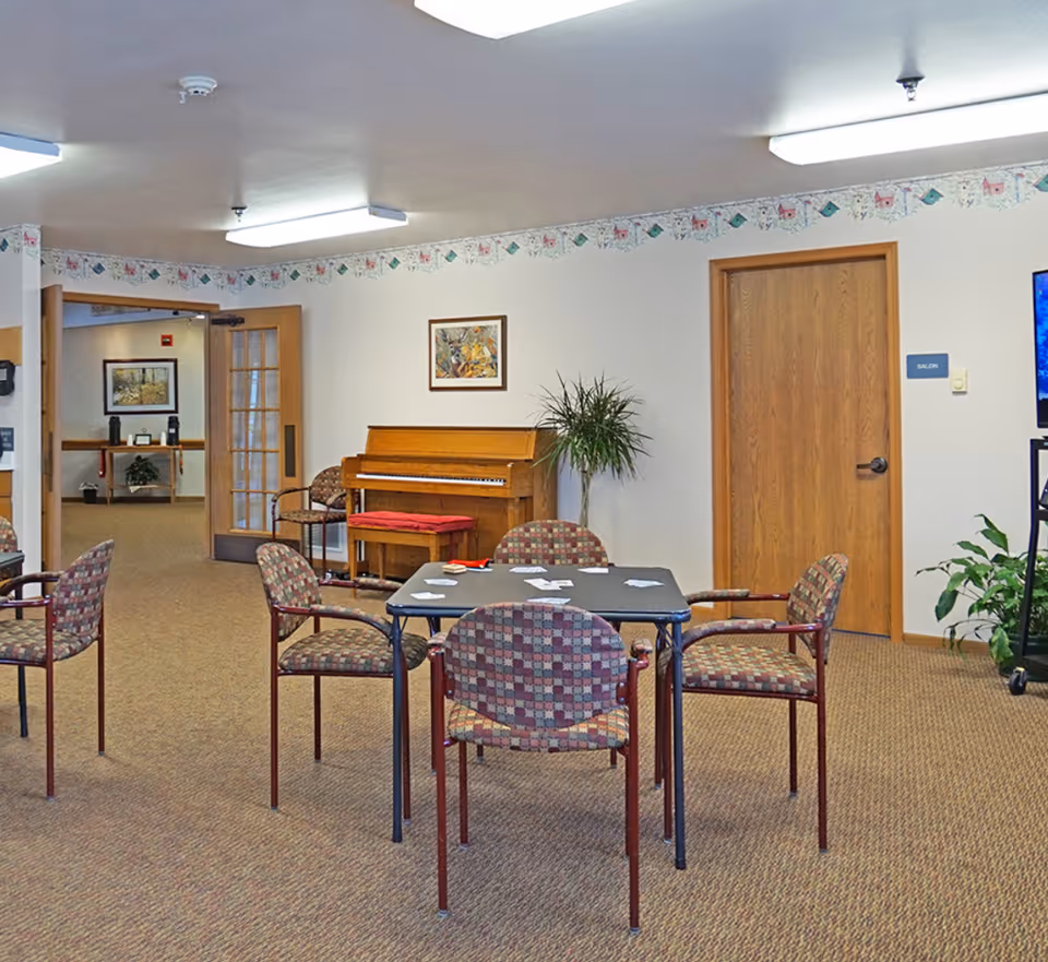 A common area in Pines Assisted Living featuring a square table with four patterned chairs around it. On the table are scattered playing cards. In the background, there is an upright piano with a red cushioned bench, a potted plant, a framed picture on the wall, and a wooden door labeled 'Salon'. The room has beige carpeting and fluorescent ceiling lights.
