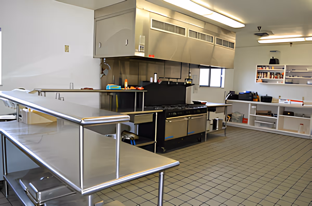 A clean, industrial kitchen with stainless steel counters and appliances, including a large stove with an overhead vent. Shelves and storage units are visible along the walls, with various kitchen supplies and equipment placed on them. The floor is tiled, and the room is well-lit with fluorescent lights.