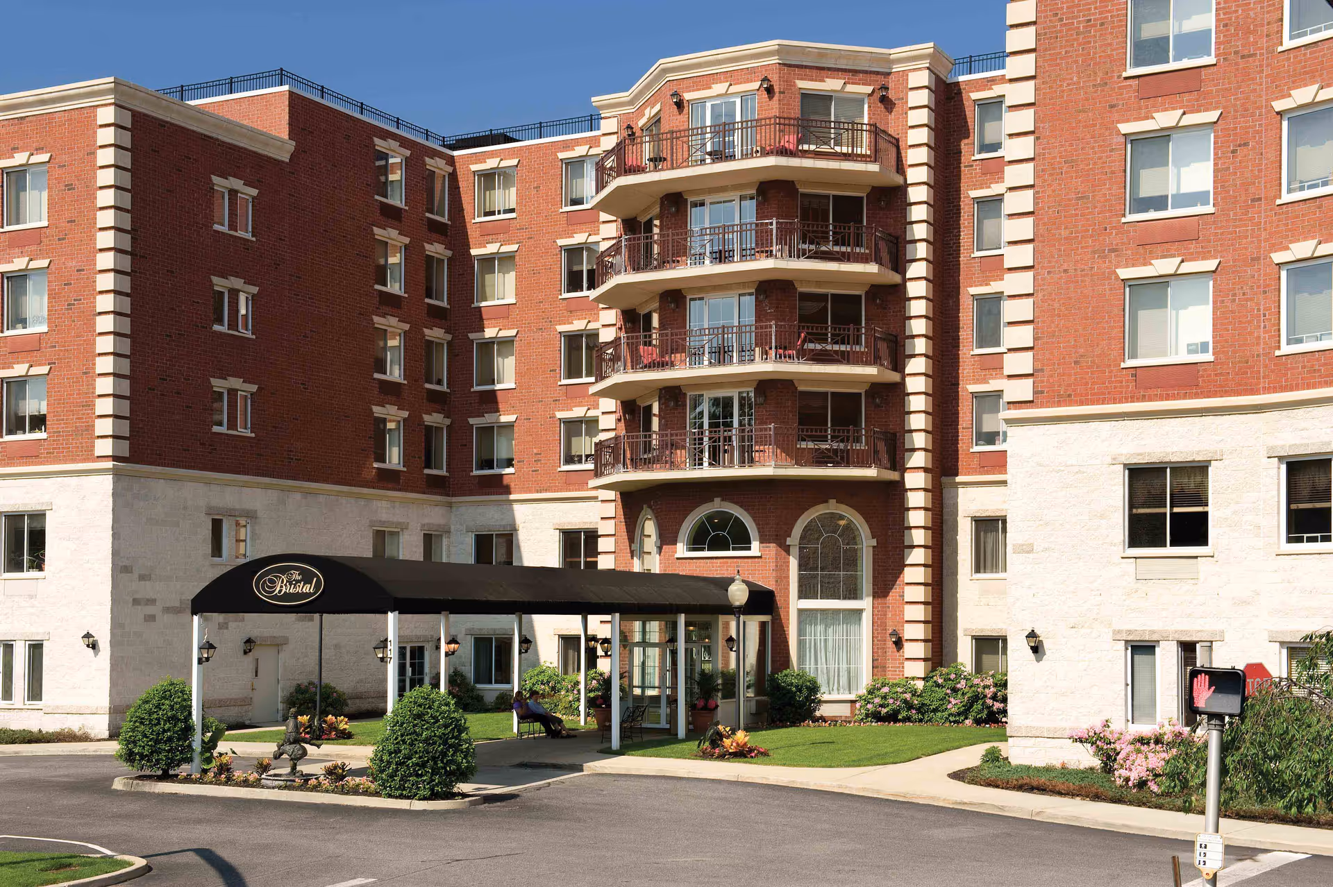 Exterior view of The Bristal Assisted Living at Westbury building with red brick and white stone facade, multiple balconies, a covered entrance with black canopy, and landscaped greenery around the entrance.