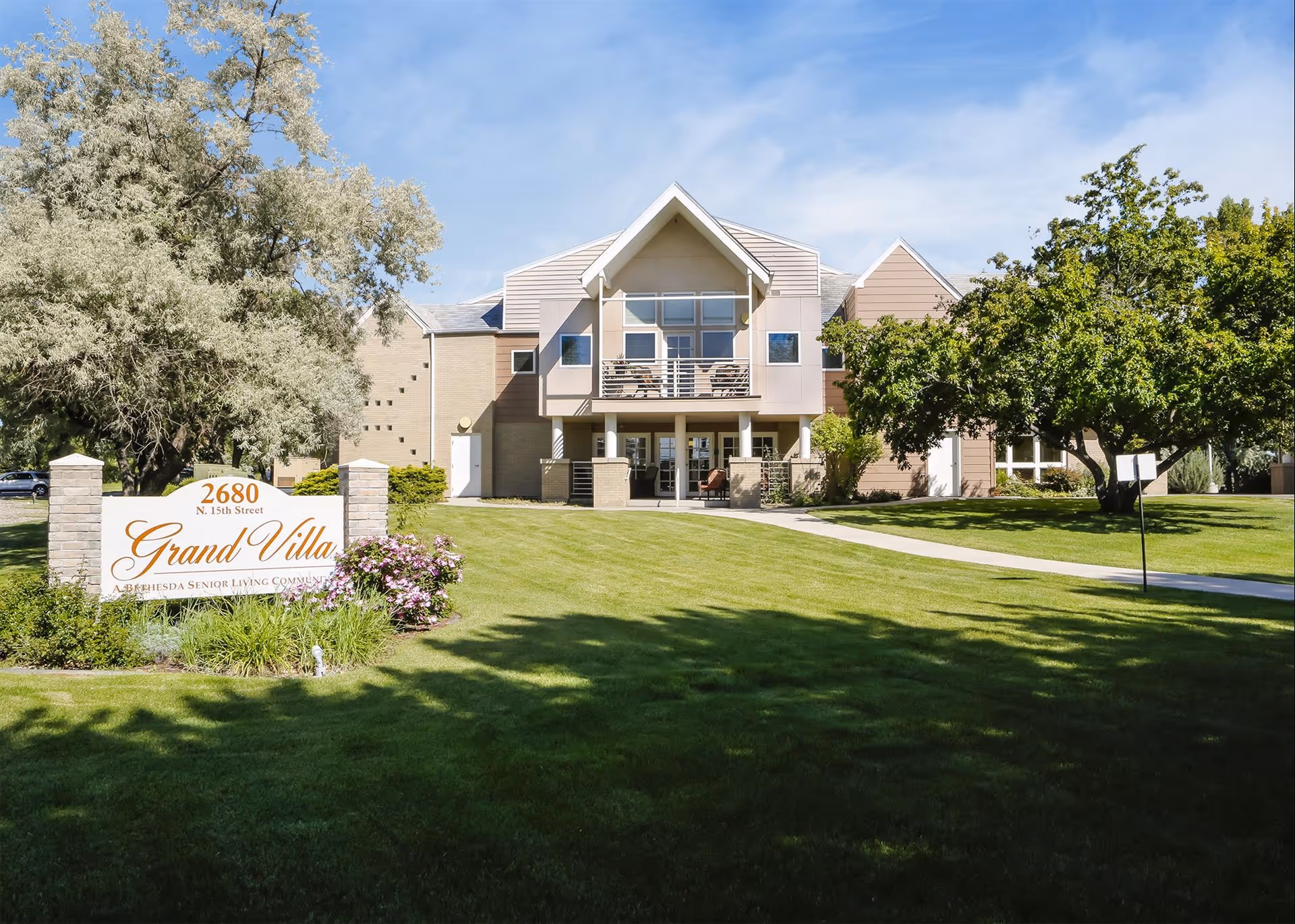 Front exterior of the Grand Villa assisted living building with a manicured lawn, trees, walkway, and a sign in the foreground.