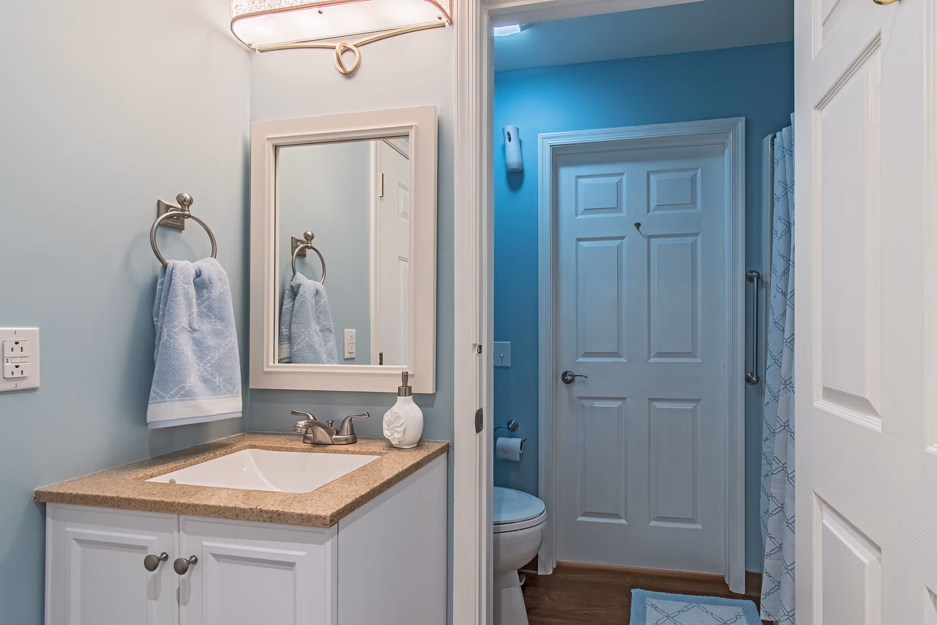 Small bathroom with a vanity and sink, mirror and towel, and a toilet visible through an open door.