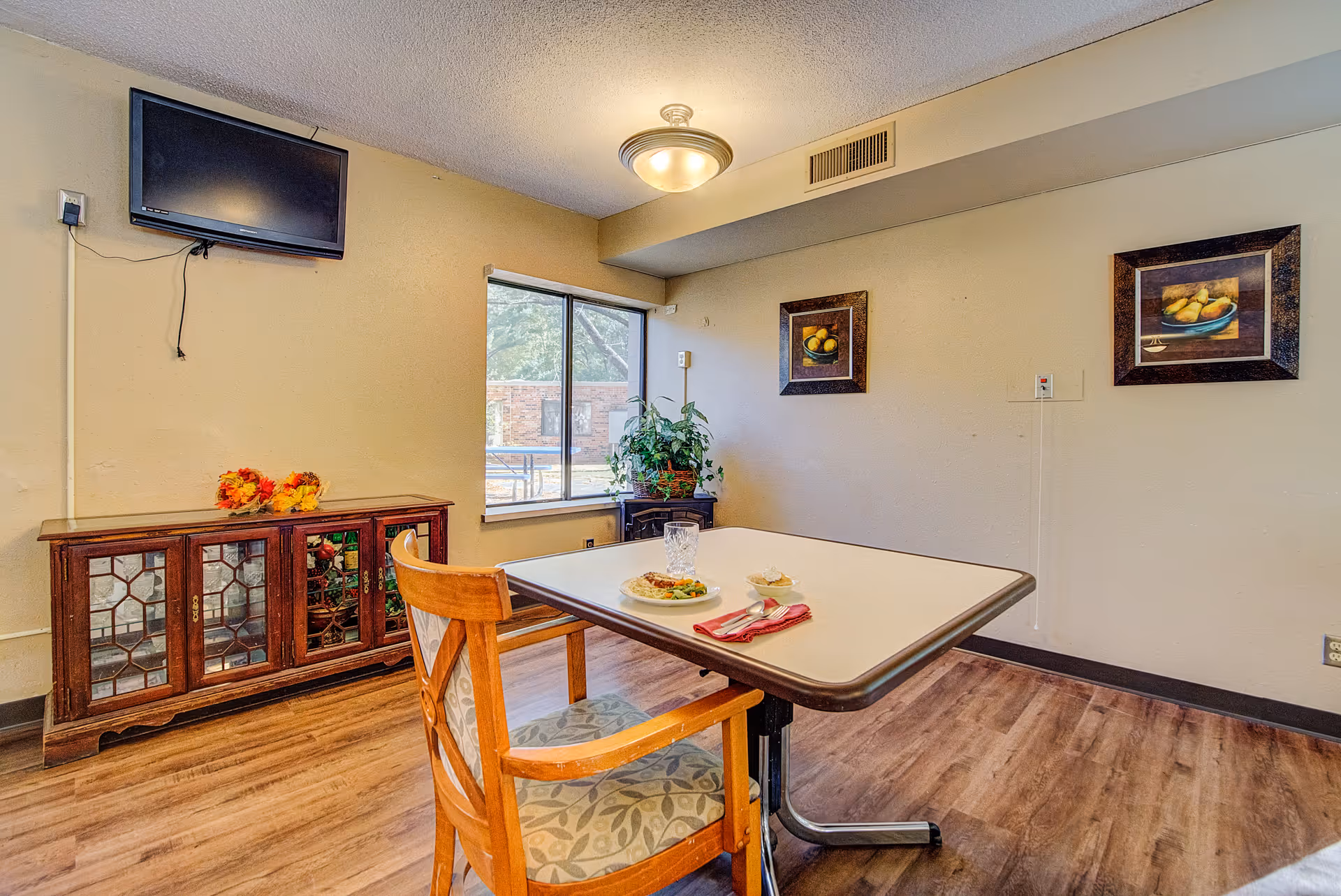 A small dining area with a square table set for one with a plate of food, a glass, and a napkin with utensils. There is a wooden chair with a patterned cushion. The room has wood flooring, a window showing an outdoor view, a wooden cabinet with glass doors, two framed fruit paintings on the wall, and a wall-mounted TV.