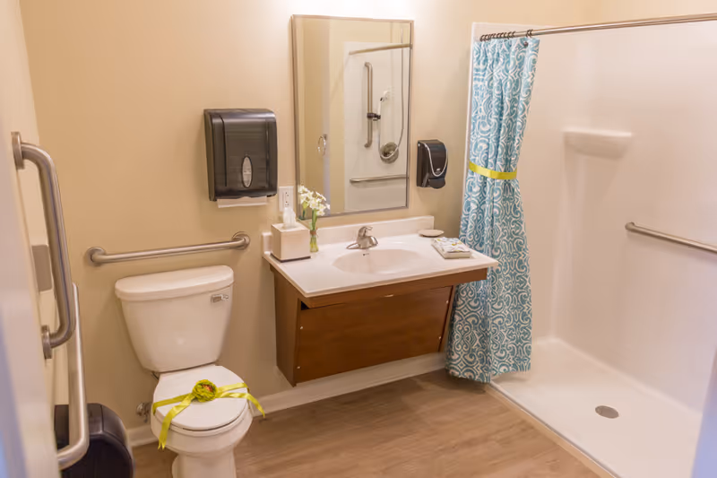 A clean and accessible bathroom featuring a white toilet with a green ribbon decoration, a wall-mounted sink with a mirror above it, a tissue box, a small vase with white flowers, and a soap dish. There is a walk-in shower with a blue patterned shower curtain tied back with a yellow ribbon, grab bars on the walls, and a wooden floor.