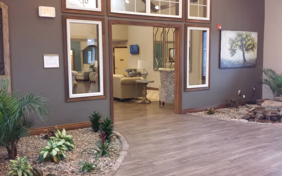 Indoor area of a senior living facility with a wooden floor, a small indoor garden with plants and rocks on the left and right sides, and a seating area with beige sofas and armchairs visible through a large open doorway. The walls are painted gray and decorated with mirrors and a painting of a tree. An exit sign is visible above the doorway.