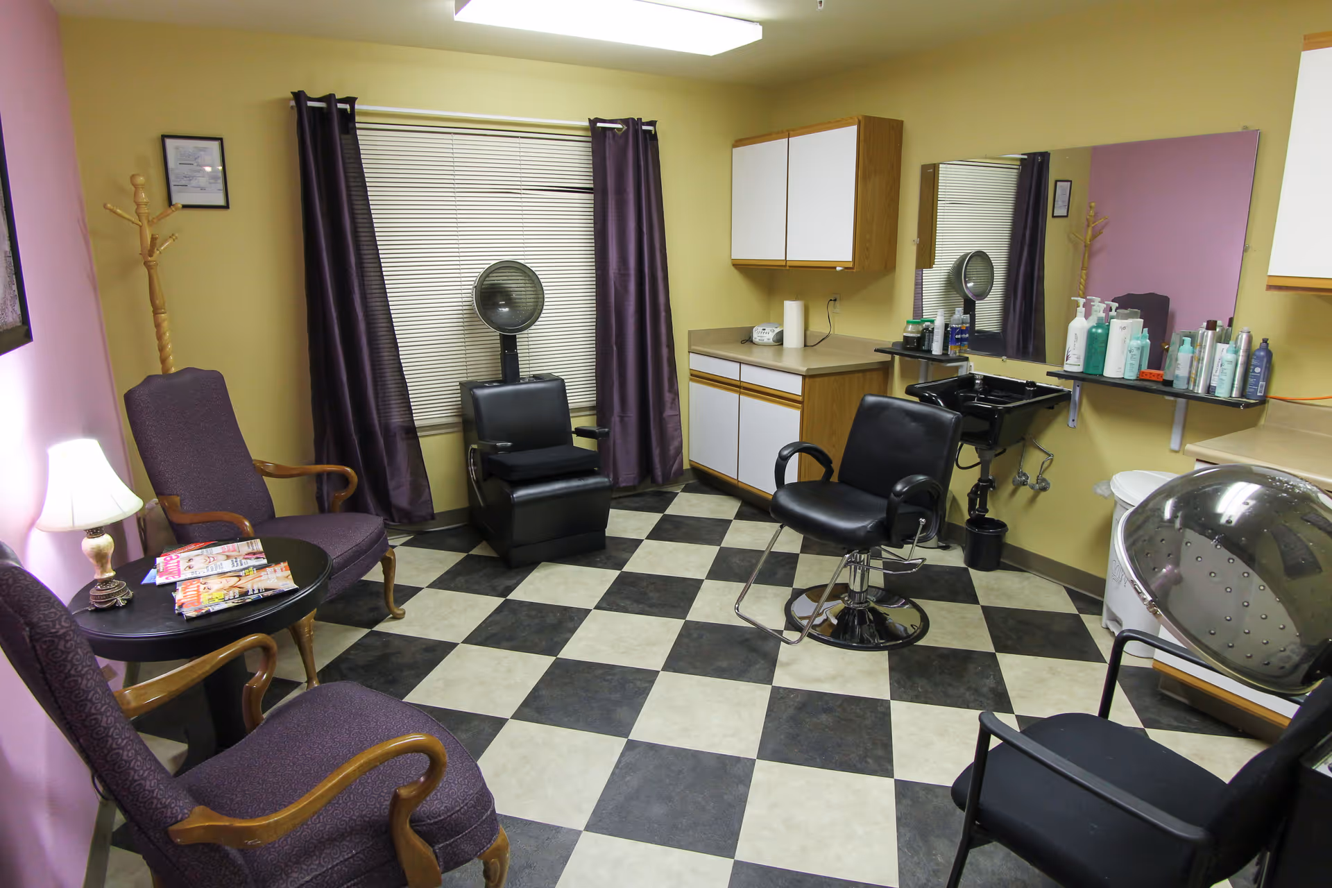 Interior of a hair salon room with checkered black and white floor tiles, two purple armchairs with wooden arms around a small round table with magazines, a black salon chair in front of a mirror and sink, a hair dryer chair, and cabinets along the walls. The walls are painted yellow and purple with a window covered by blinds and purple curtains.