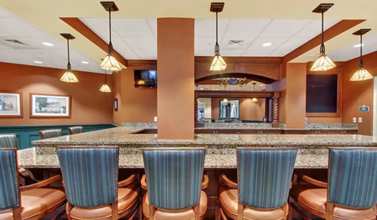 Interior view of a senior living facility dining area with a granite countertop bar surrounded by blue striped cushioned chairs. The room features warm brown walls, hanging pendant lights, framed artwork, and a mounted television.