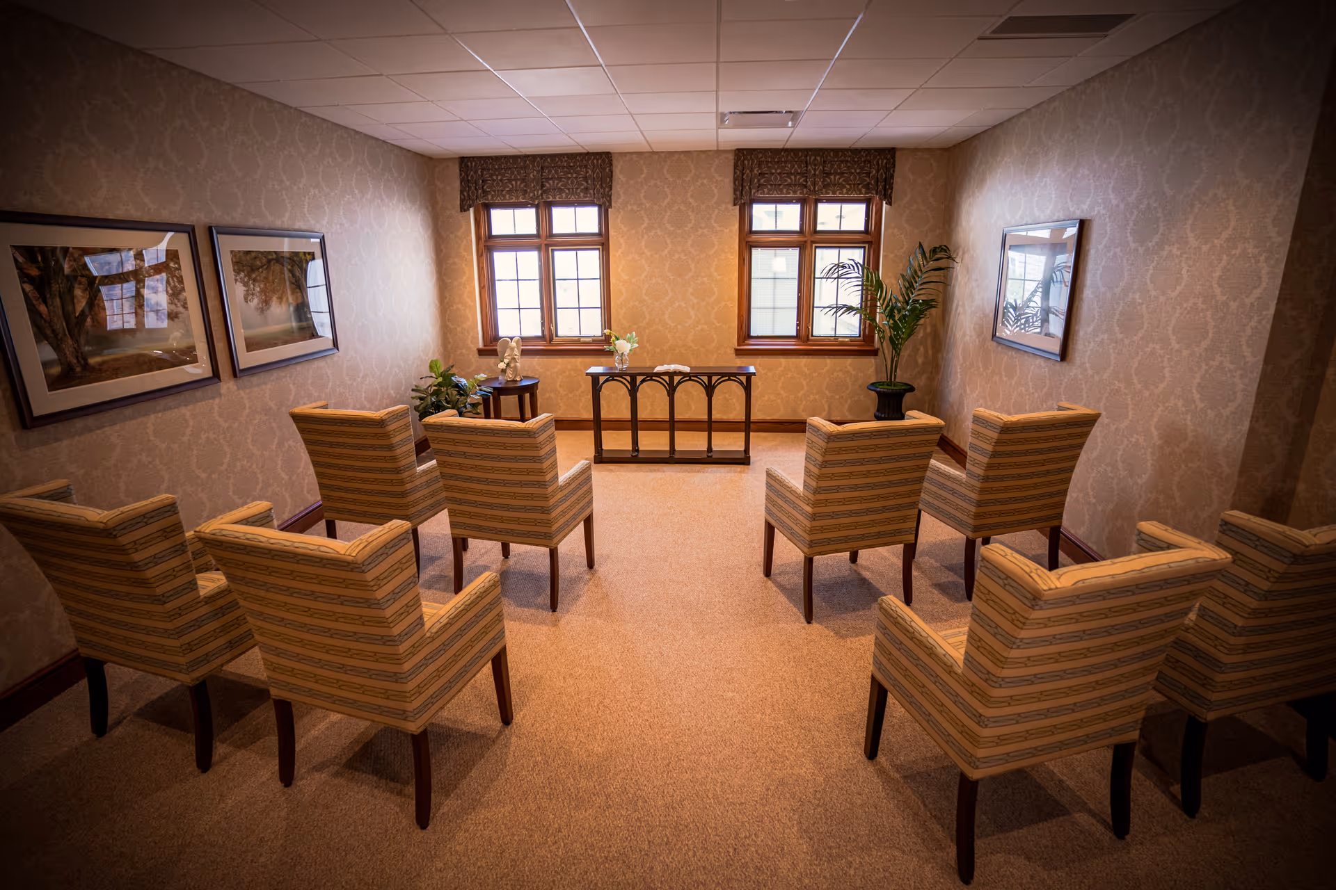 Small meeting room with upholstered armchairs arranged in rows facing a console table under two windows in a warmly decorated interior.