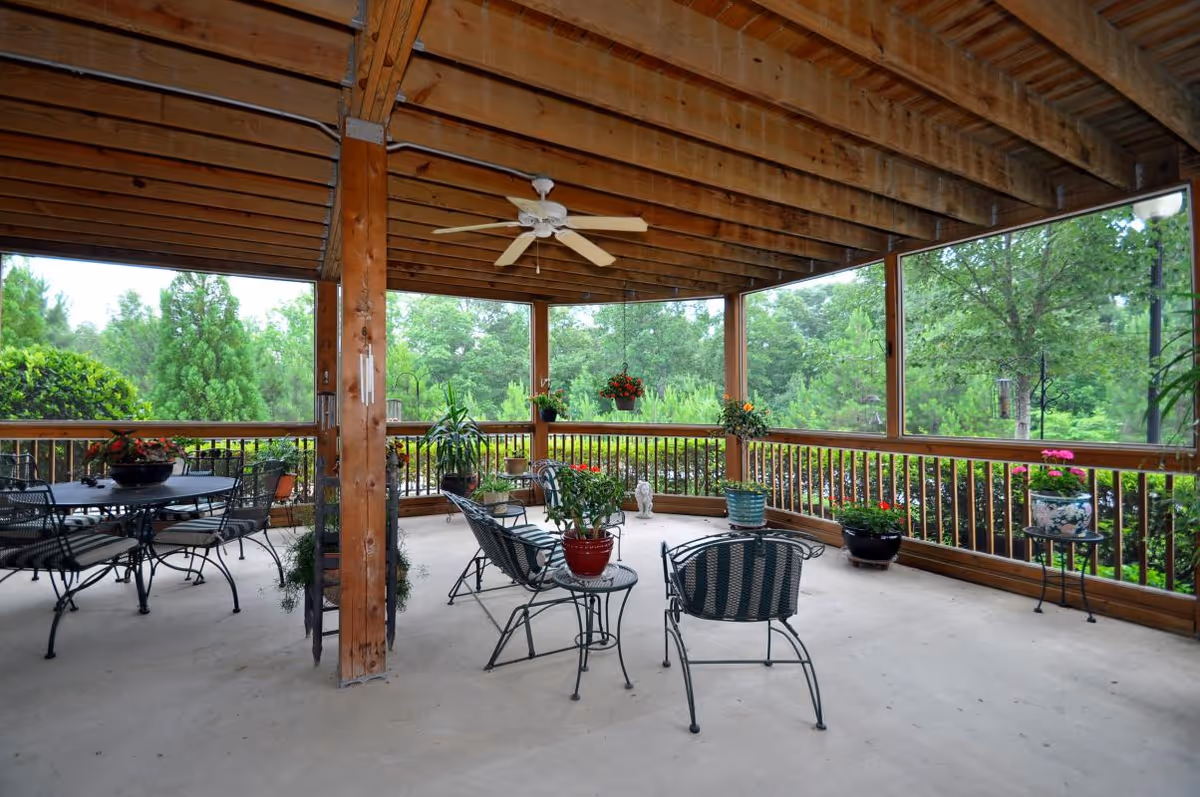 Covered wooden patio with a ceiling fan, metal tables and chairs, and potted plants overlooking a green landscape.