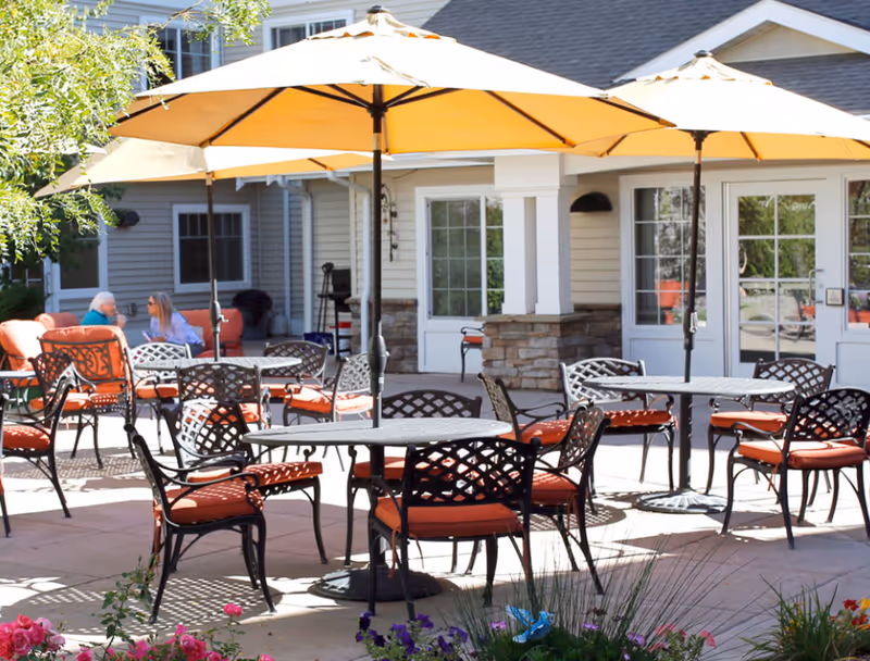 Outdoor patio area with several round metal tables and chairs with orange cushions, shaded by large yellow umbrellas. In the background, two people are sitting and talking on an orange cushioned bench near a building with white doors and windows. There are flowers and greenery in the foreground.