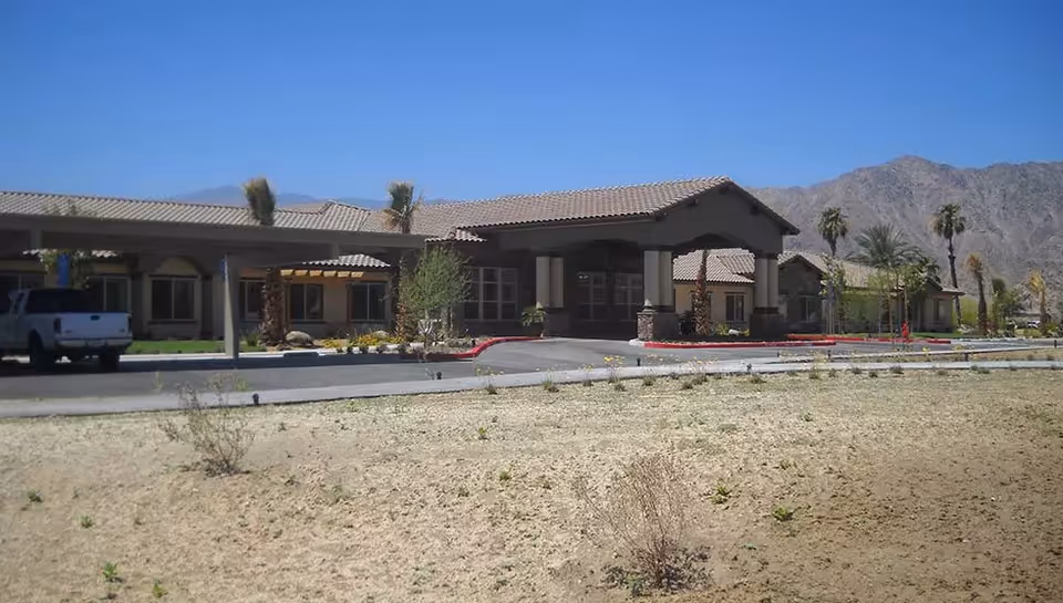 Exterior view of a single-story assisted living facility with a covered entrance, beige walls, and a tiled roof. There is a driveway with a white pickup truck parked on the left side. The landscape in front is dry with sparse vegetation, and mountains are visible in the background under a clear blue sky.