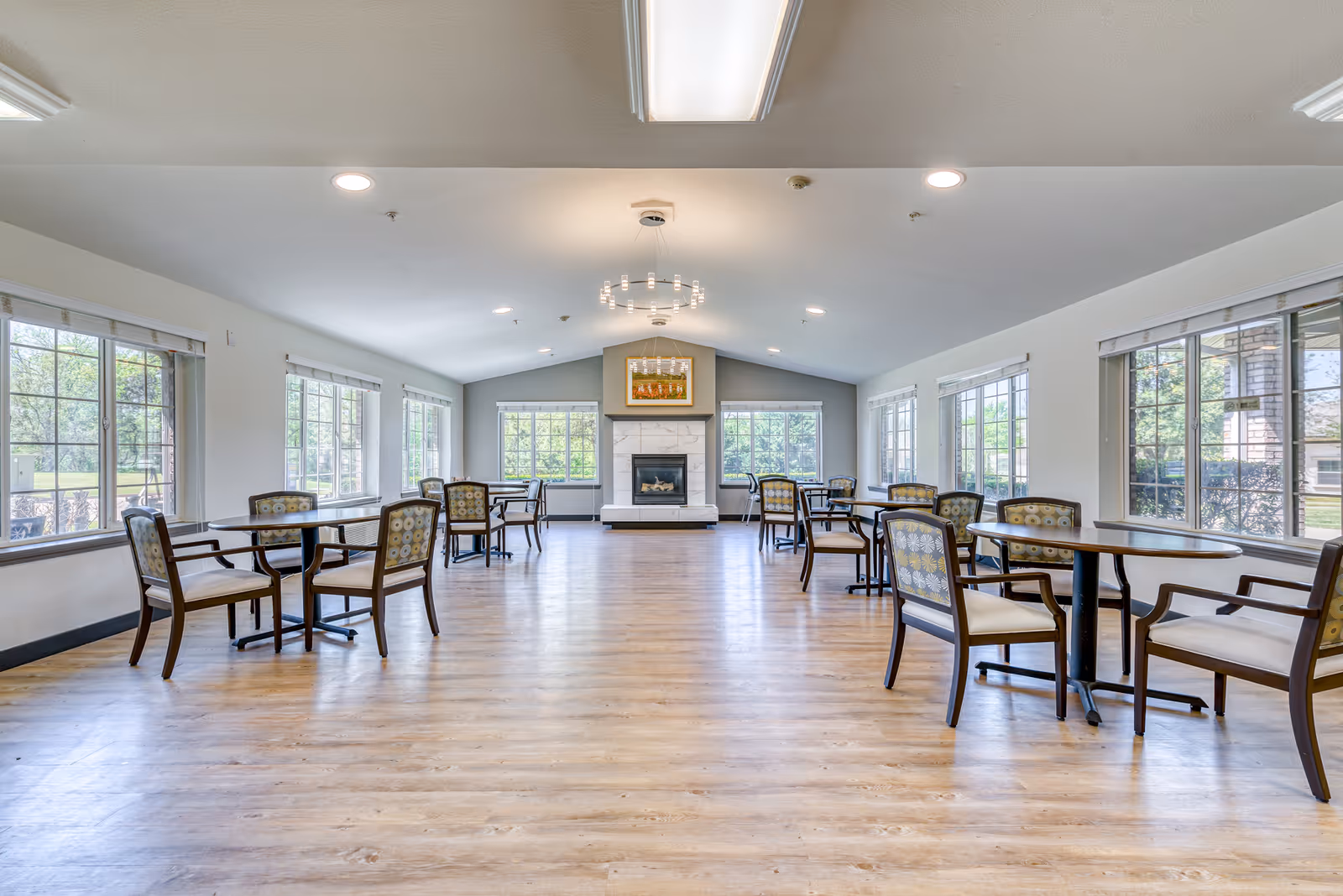 A spacious dining room with multiple round tables and chairs arranged neatly. Large windows on both sides allow natural light to fill the room. At the far end, there is a fireplace with a framed picture above it. The floor is wooden, and the ceiling has recessed lighting and a modern chandelier.