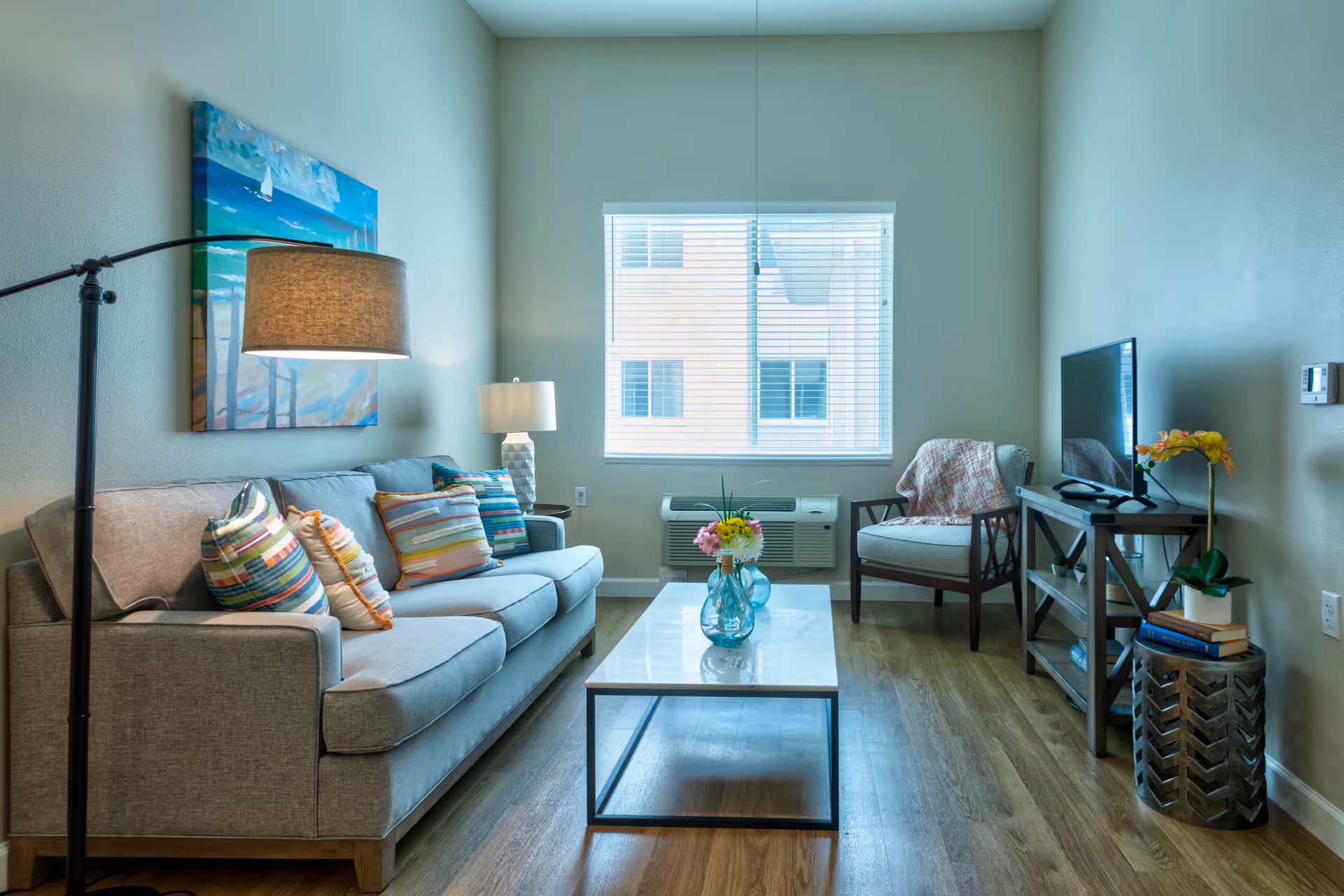 A bright living room with a gray sofa adorned with colorful striped pillows, a floor lamp, a coffee table with a blue vase holding flowers, a single armchair with a throw blanket, a TV on a wooden stand, and a window with blinds letting in natural light.