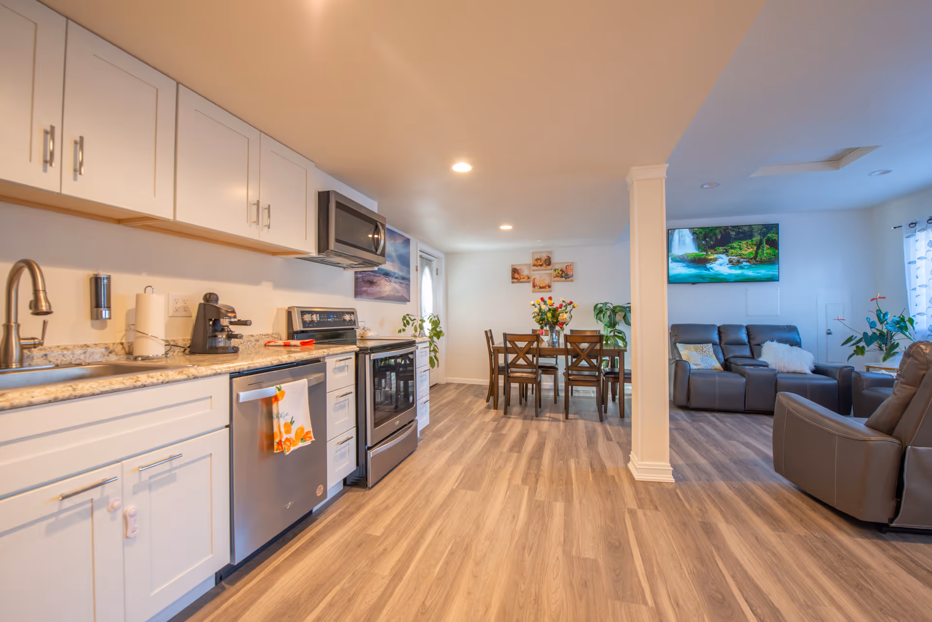 Open-plan modern kitchen with white cabinets and stainless steel appliances leading to a dining table and a living area with recliner sofas.