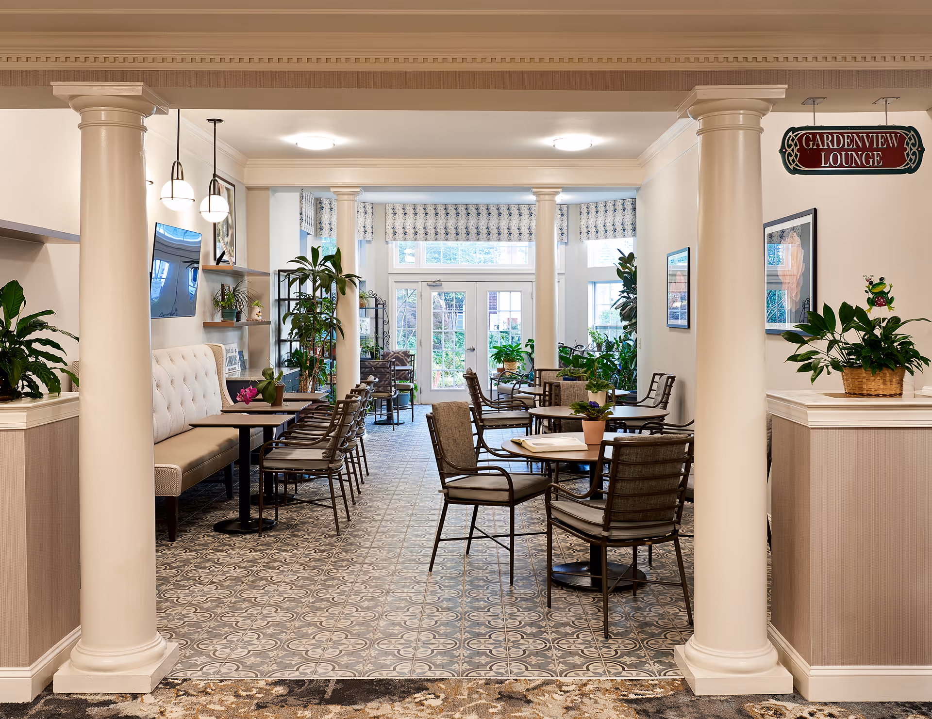 A bright and inviting lounge area with patterned tile flooring, round tables with chairs, cushioned bench seating along one wall, and several potted plants. The space is framed by two large white columns and has large windows and a glass door at the far end letting in natural light. A sign hanging from the ceiling reads 'Gardenview Lounge'.