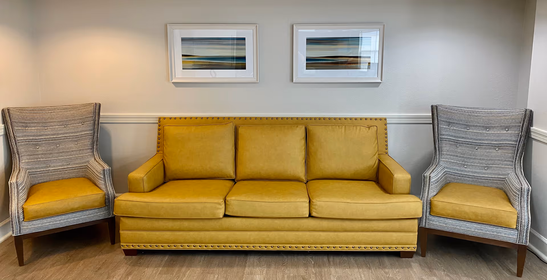 A seating area with a mustard yellow three-seat sofa flanked by two gray armchairs with mustard yellow cushions, set against a light-colored wall with two framed abstract landscape paintings above.