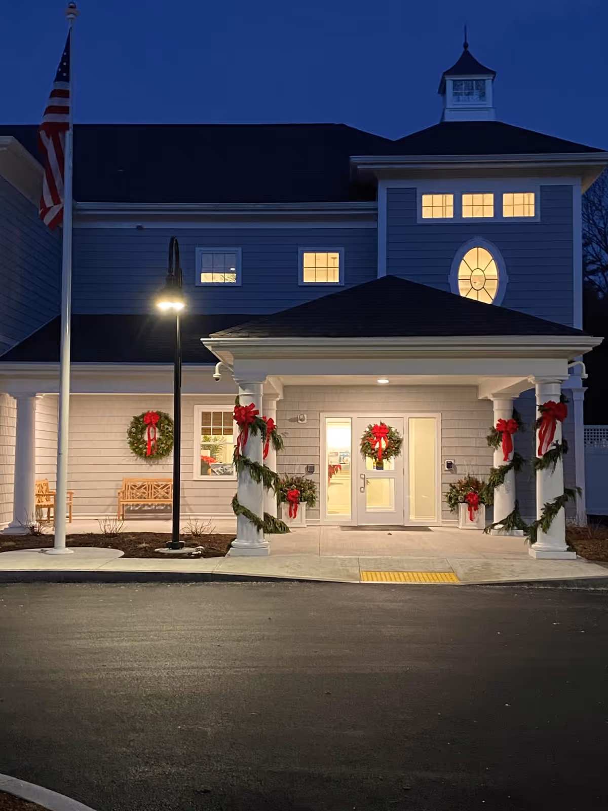 Exterior front entrance of a healthcare and retirement facility at dusk, decorated with Christmas wreaths and garlands with red bows on white columns and doors. An American flag is on a pole to the left, and warm lights glow from the windows.
