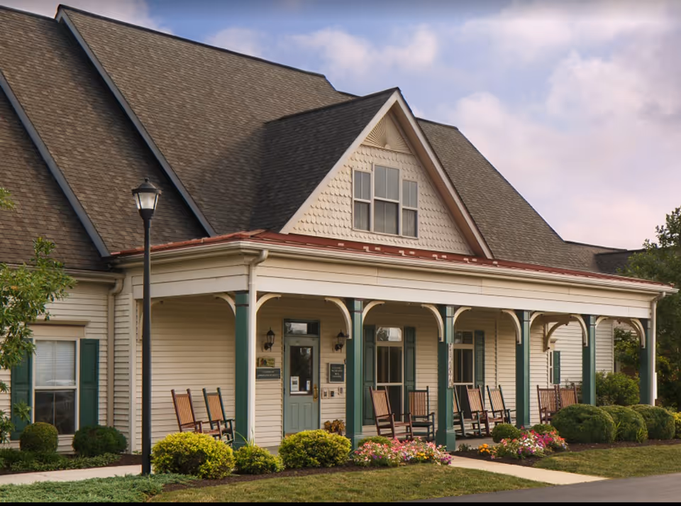 Exterior view of Columbia Cottage Linglestown showing a building with a large porch featuring several wooden rocking chairs. The building has beige siding, green window shutters, and a steeply pitched roof. There are bushes and flowers planted along the front of the porch and a lamppost near the sidewalk.