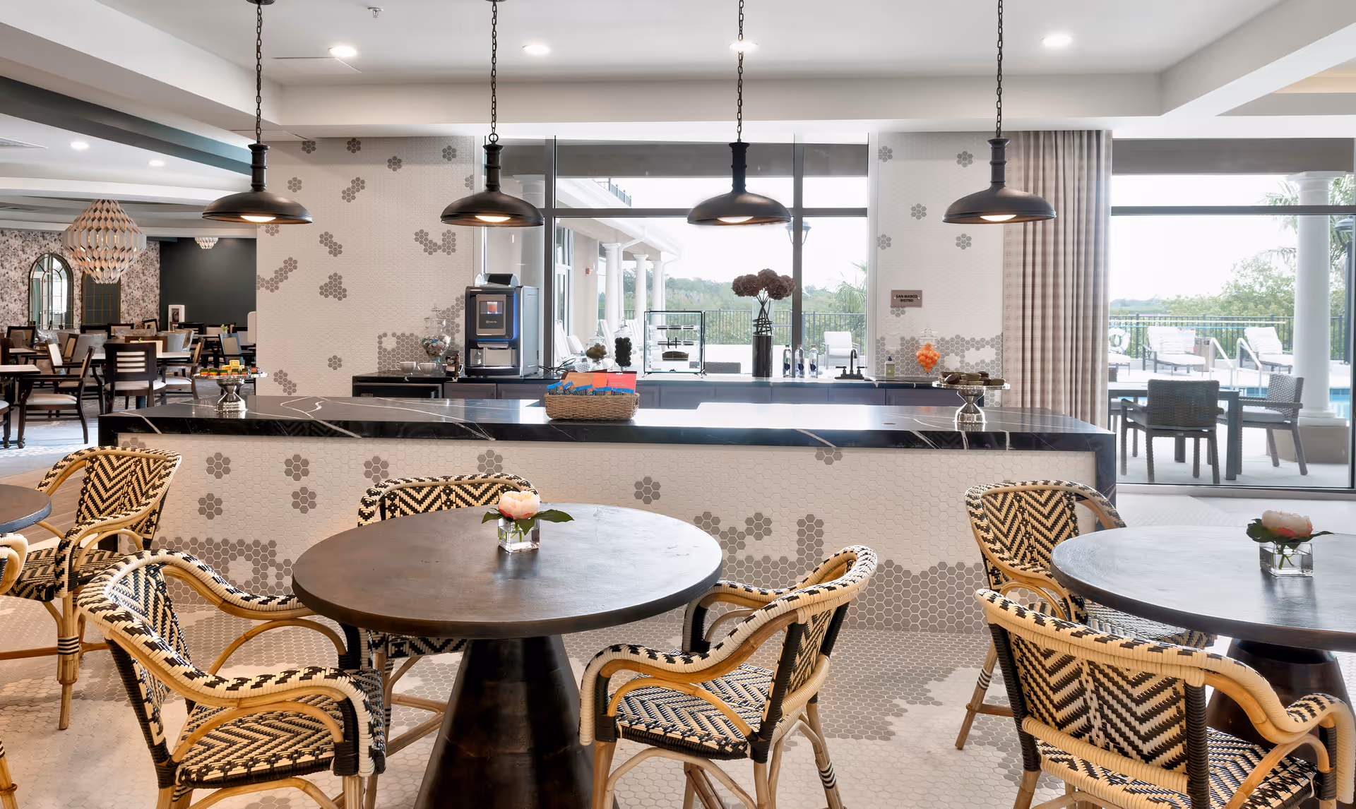 A bright and modern dining area in an assisted living facility featuring round tables with woven chairs. The room has a counter with a coffee machine and decorative items, pendant lights hanging from the ceiling, and large windows offering a view of an outdoor patio with seating and greenery.