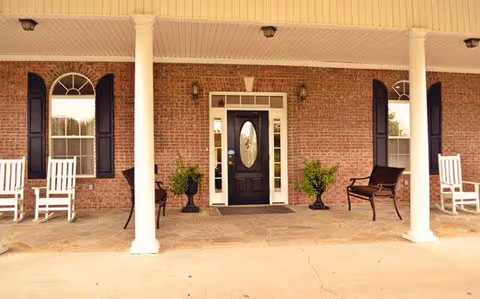 Front porch of a brick building with a black door featuring an oval glass panel, flanked by two windows with black shutters. The porch has white columns, two white rocking chairs on the left, a black bench on the right, and two potted plants near the entrance.