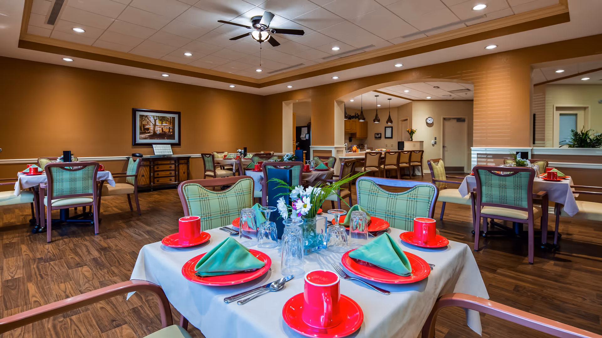 A dining room in a senior living facility with tables set for a meal. Each table has red plates and cups, green folded napkins, silverware, and a small floral centerpiece. The room has wooden flooring, beige walls, and a ceiling fan. In the background, there is a kitchen area with bar stools and pendant lights.