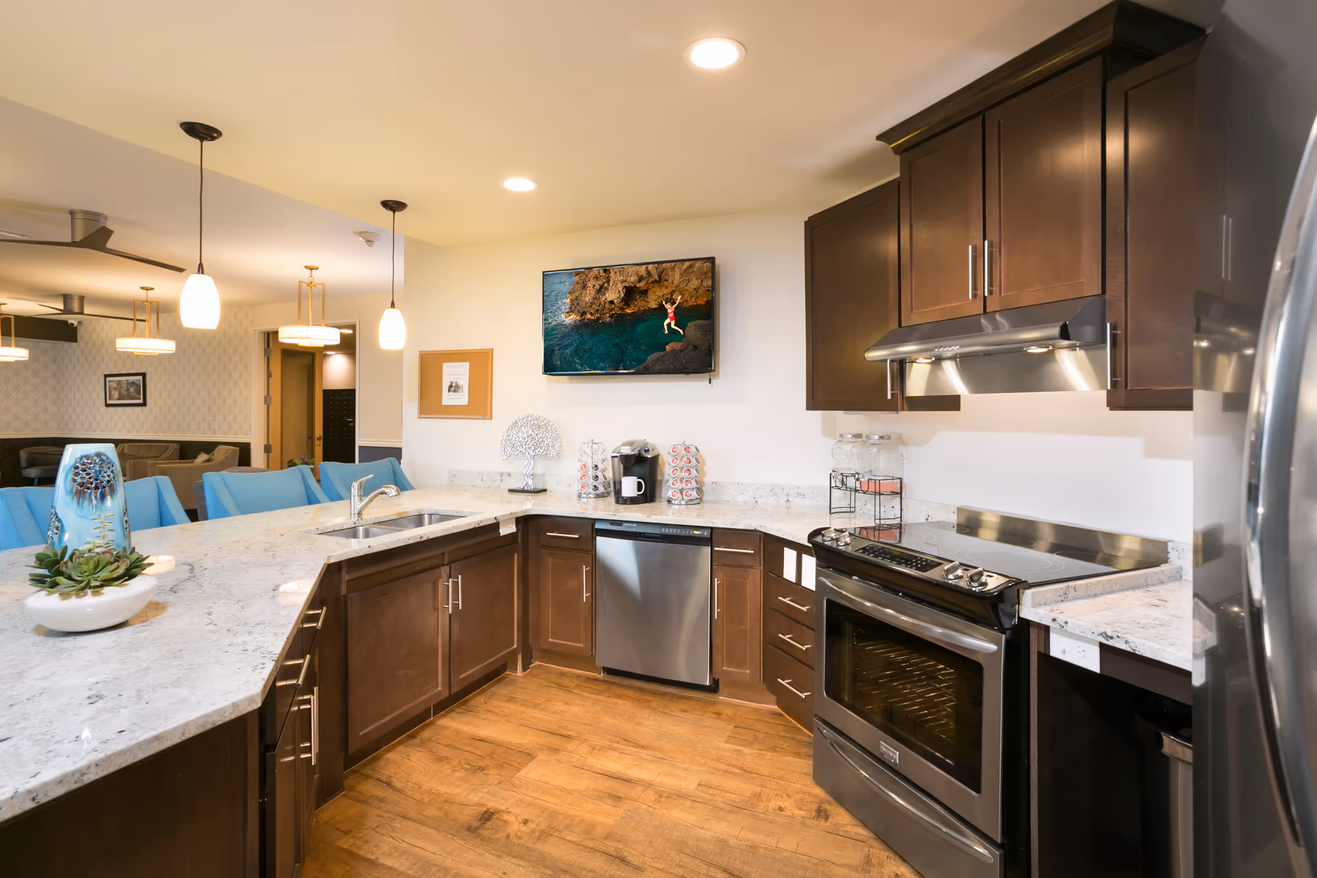 Modern kitchen area with dark wood cabinets, stainless steel appliances including an oven, dishwasher, and refrigerator. Light granite countertops with a sink and decorative items. Pendant lights hang over the counter, and a flat-screen TV is mounted on the wall showing a person jumping into water from a cliff. In the background, a seating area with blue chairs and additional lighting fixtures is visible.