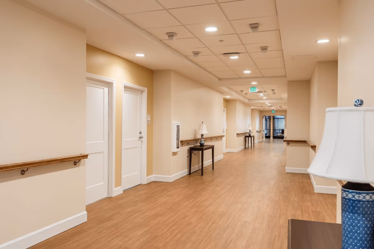 A long, well-lit hallway in a senior living facility with light beige walls, wooden handrails, and wood-look flooring. There are white doors along the left side and small tables with lamps placed against the walls. The ceiling has recessed lighting and an exit sign is visible at the far end of the hallway.
