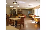 A spacious dining room with multiple tables and chairs arranged neatly. The room is well-lit with ceiling lights and natural light coming through large windows with curtains. The floor is wooden, and the walls are painted in a light color.