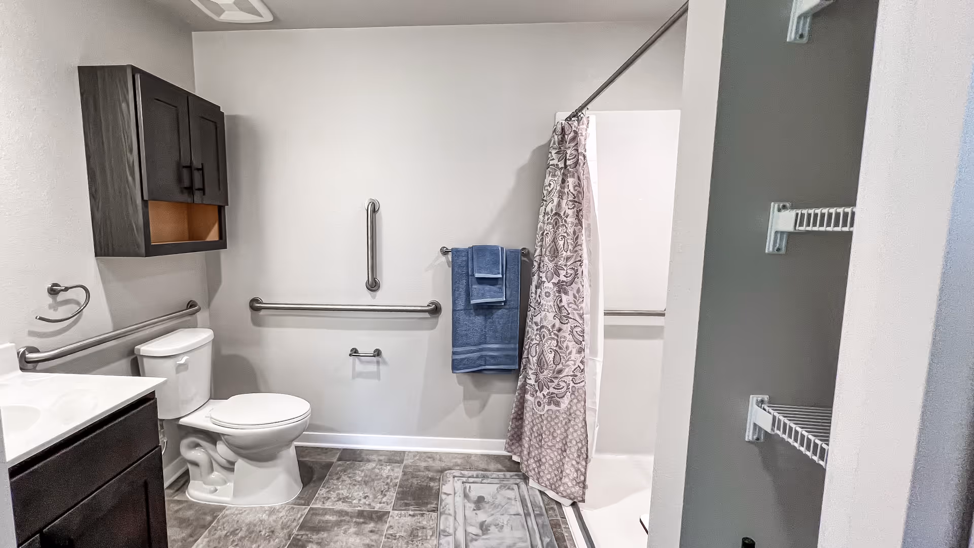 A clean and accessible bathroom featuring a white toilet, a dark wood cabinet above it, a sink with a dark wood vanity, and a shower with a patterned curtain. There are multiple stainless steel grab bars on the walls for support, a blue towel hanging on a towel rack, and a gray bath mat on the tiled floor.