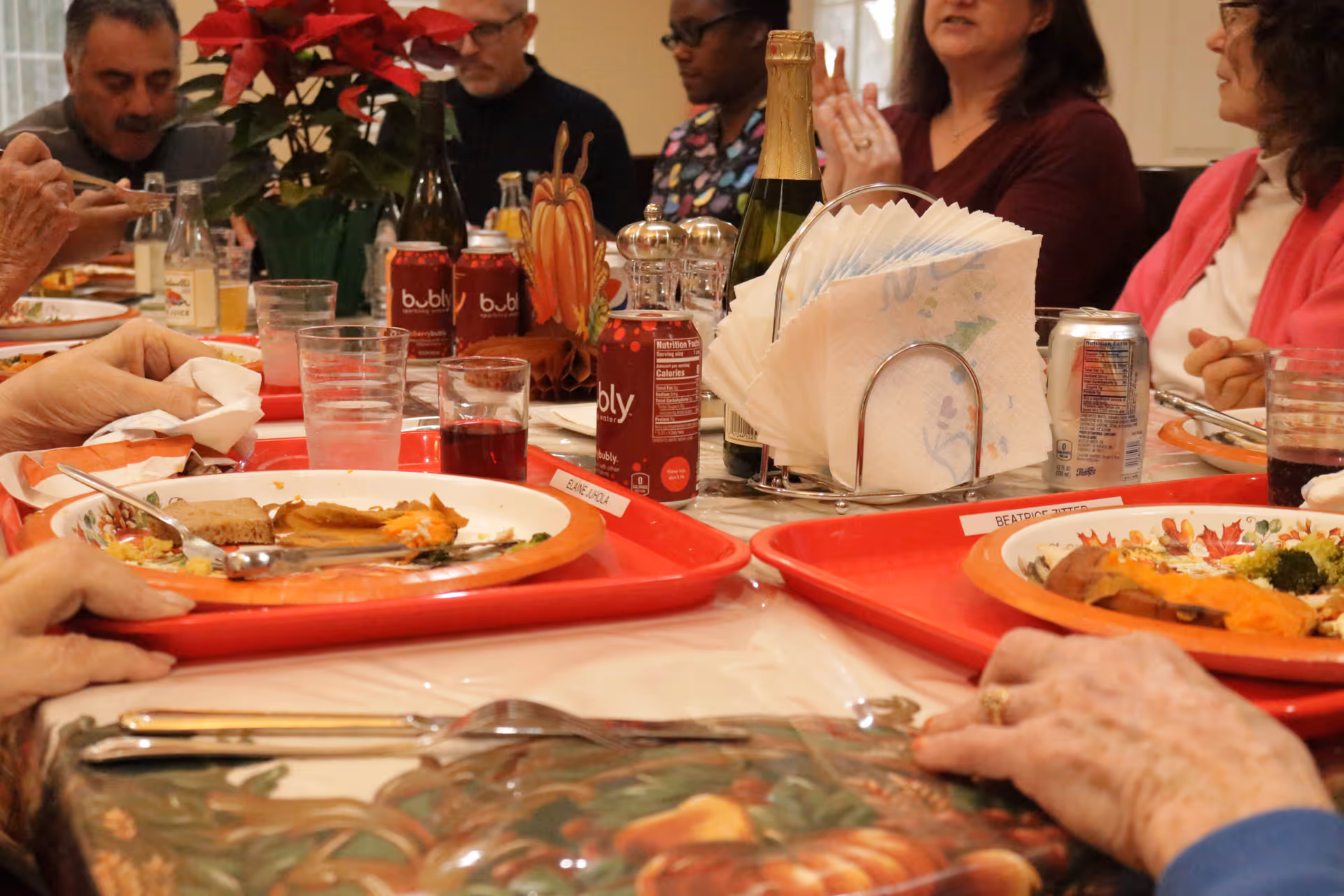 A group of people sitting around a table enjoying a meal together with red trays holding plates of food, drinks, and napkins. The table is decorated with a fall-themed tablecloth and a centerpiece featuring a poinsettia and a small pumpkin decoration.