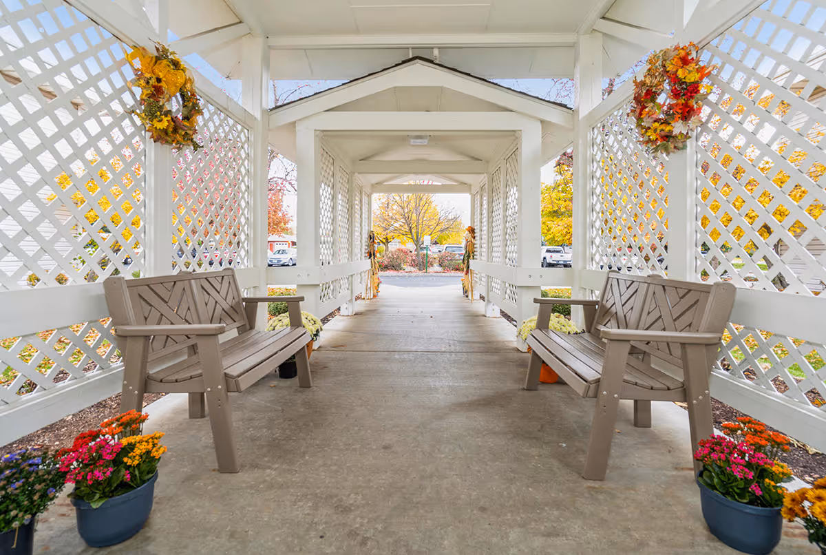 Covered outdoor walkway with white lattice walls on both sides, decorated with autumn wreaths and potted flowers. Two brown benches face each other along the walkway, and trees with fall foliage are visible in the background.