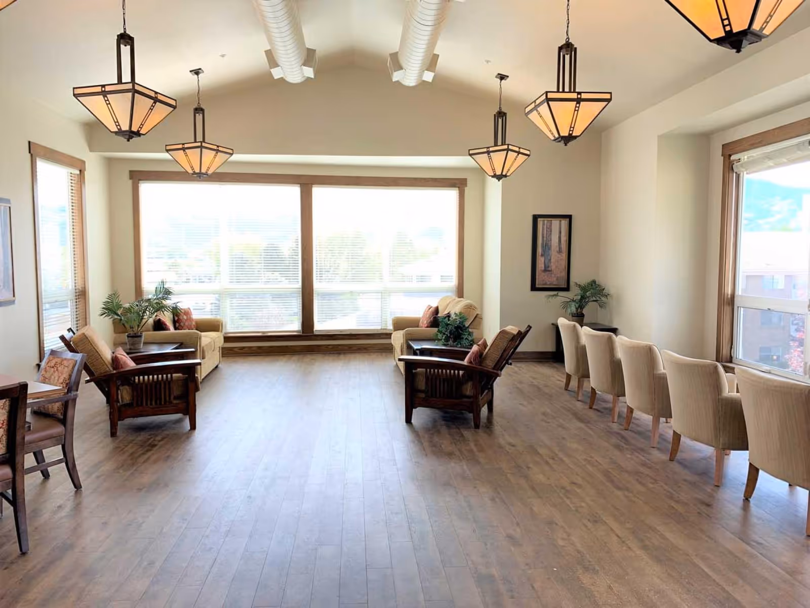 Bright communal lounge with sofas, armchairs, large windows and pendant lights in an assisted living facility.