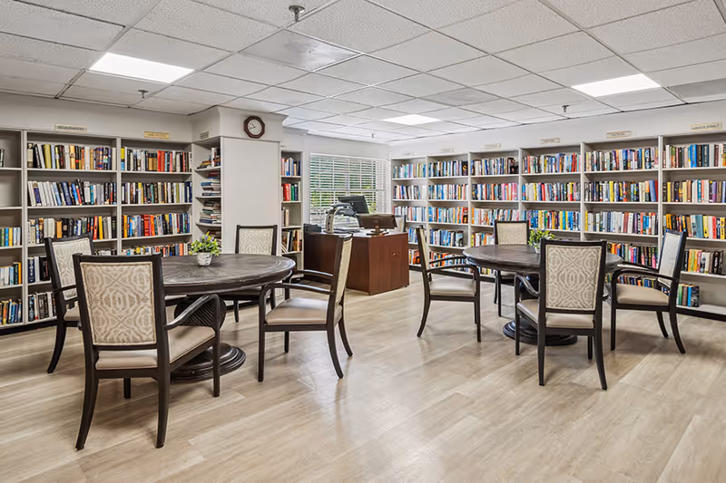 A bright and spacious library room with light wood flooring, two round tables each surrounded by four upholstered chairs, and walls lined with white bookshelves filled with books. A wooden desk with a computer is positioned near a window with white blinds, and a clock is mounted on a column in the center of the room.