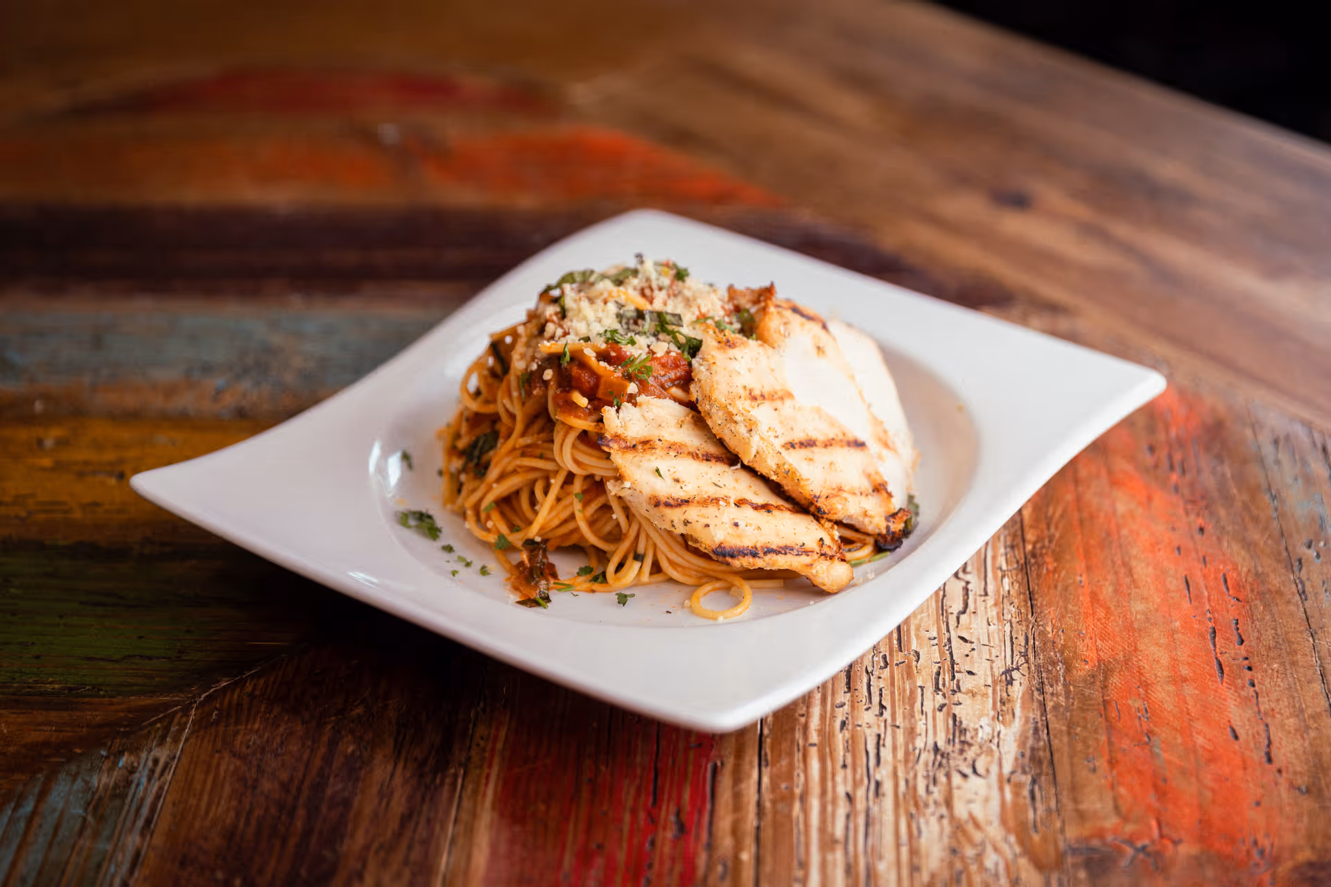 A white square plate with grilled chicken breast slices served on a bed of spaghetti pasta topped with tomato sauce and garnished with herbs and grated cheese, placed on a rustic wooden table.