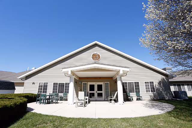 Exterior view of a single-story building with beige siding and a peaked roof under a clear blue sky. The building has a covered entrance supported by two white columns, with several green chairs and tables arranged on the concrete patio in front. There is a tree with white blossoms on the right side and trimmed bushes on the left.