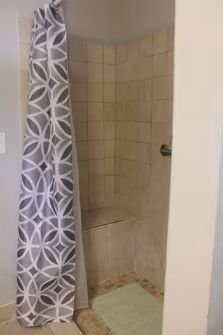A tiled shower area with beige square tiles on the walls and a small built-in bench. There is a gray and white patterned shower curtain partially pulled to the side, and a green bath mat on the floor outside the shower.