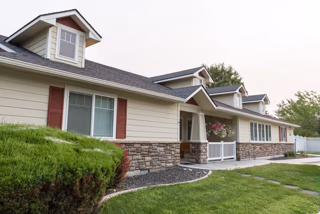 Exterior view of a single-story residential building with beige siding, stone accents, and red shutters. The building has multiple dormer windows on the roof and a covered porch with hanging flower baskets. There is a well-maintained lawn and shrubbery in the foreground.
