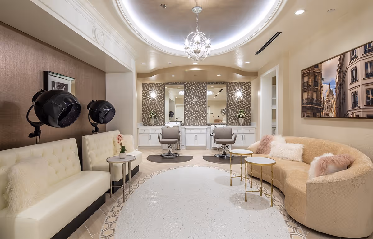 A stylish salon room with two hair drying chairs on the left, two salon chairs in front of mirrors with patterned tile walls, and a curved beige couch with fluffy pillows on the right. The room has a soft, warm lighting with a decorative ceiling light fixture and a large photo of a cityscape on the wall.