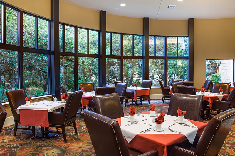 A dining room with multiple tables covered in white and red tablecloths, each set with cups, utensils, and red folded napkins. The room features large windows with views of green trees outside and has brown leather chairs around the tables.