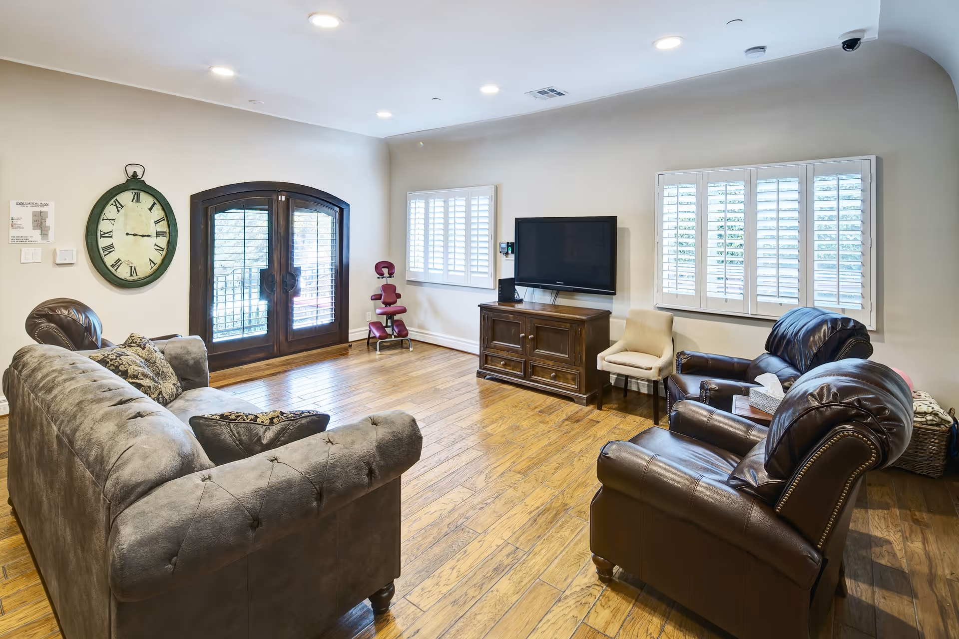 A cozy living room with wooden flooring, a gray tufted sofa, two brown leather armchairs, a wooden TV stand with a flat-screen TV mounted on the wall, a beige chair, and a large round wall clock. The room has two windows with white shutters and double wooden doors with glass panels.
