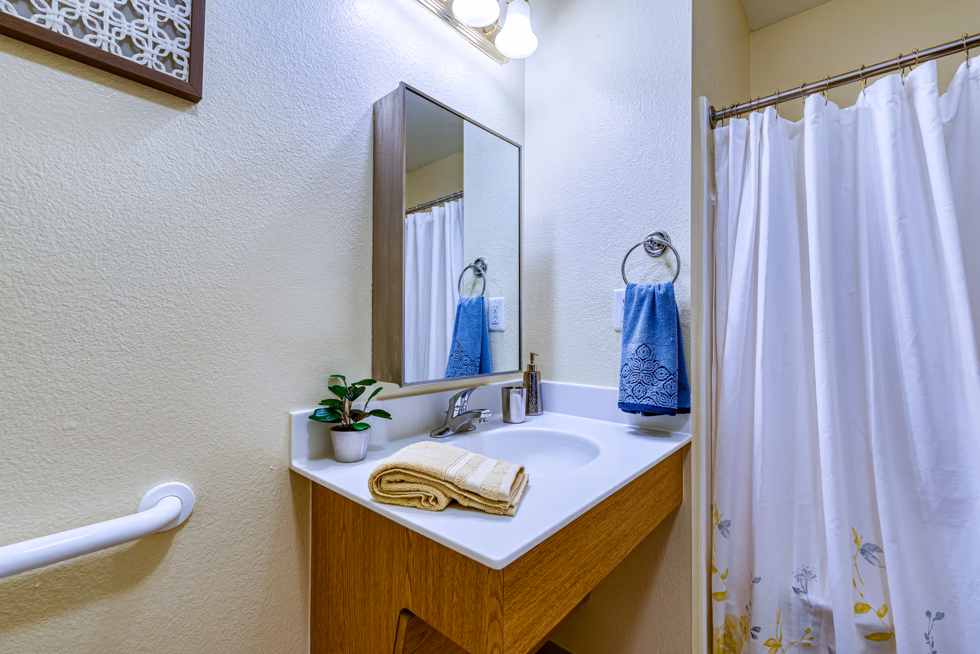 A bathroom vanity with a wooden cabinet, white countertop, and built-in sink. On the countertop, there is a small potted plant, folded beige towels, a soap dispenser, and a cup. Above the sink is a rectangular mirror with a wooden frame and a light fixture with three bulbs. A blue hand towel hangs on a ring to the right of the sink, and a white shower curtain with a floral pattern is visible in the background.