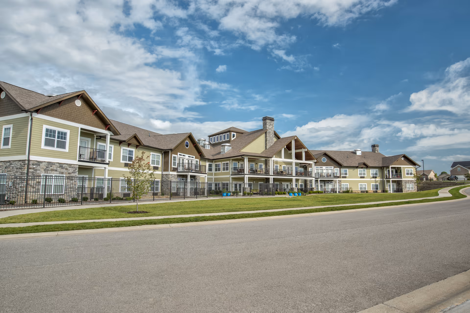 Exterior view of a large senior living facility building with multiple balconies, stone and siding facade, a fenced area, and a well-maintained lawn under a partly cloudy sky.