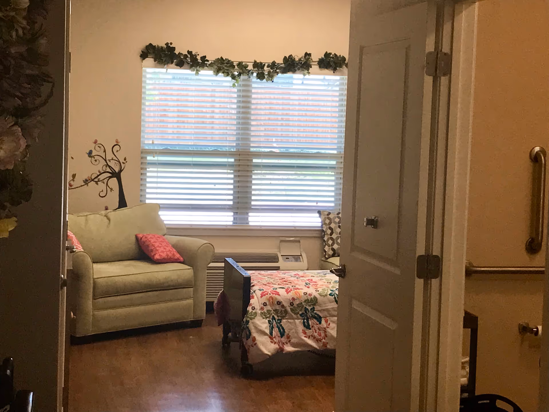 View into a cozy bedroom with a single bed covered in a floral bedspread, a light green armchair with a pink cushion, a window with blinds and a garland of greenery above it, and a partially open door revealing a bathroom with grab bars and a toilet paper holder.
